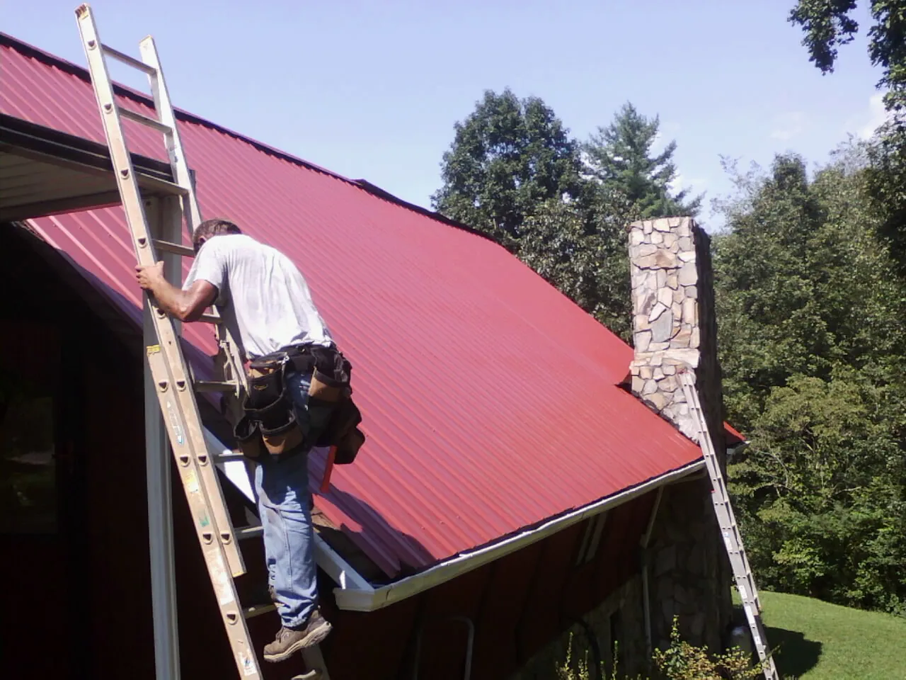 Roofer on a ladder beside a red metal roof and stone chimney — Soffit & Fascia Repair by Roofing Brothers in Washington Boro